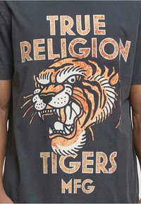 Tumši pelēks T-krekls ar lielu oranžu un melnu tigru grafiku, kas izskatās nolietojusies, un treknu baltu tekstu "TRUE RELIGION TIGERS MFG."
