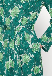 Abito floreale verde con vita con cinturino, maniche lunghe e un vivace motivo botanico su sfondo azzurro chiaro con intricate foglie.