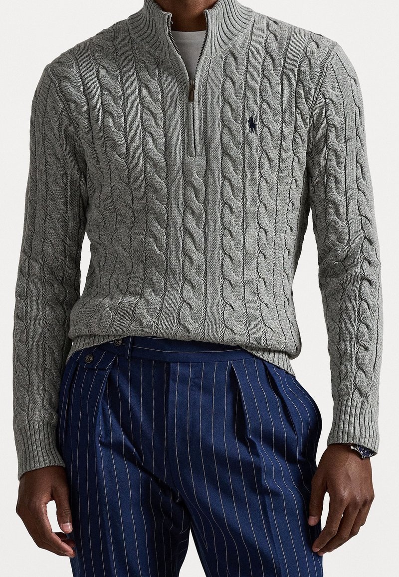 Homme portant un pull gris en maille torsadée avec zip quart et pantalon bleu marine à fines rayures, debout les bras détendus le long du corps.