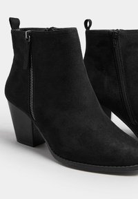 Par de botas negras de ante hasta el tobillo con tacones cuadrados y cremalleras laterales sobre un fondo blanco.
