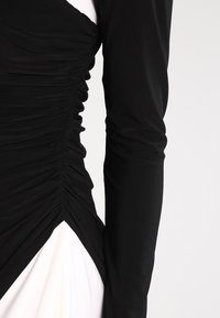 Svart klänning med långa ärmar som har texturerad ruching på ena sidan, som övergår till en solid vit panel vid fållen. Mjuk, elastisk tyg.