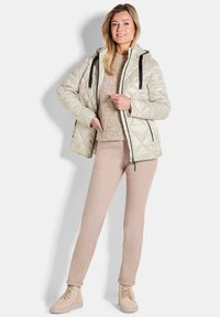 Beige gesteppte Jacke mit Kapuze, schwarzen Akzenten und Vorderreißverschluss, kombiniert mit hellrosa Hosen und beigen Schnürschuhen. Glatte Textur.