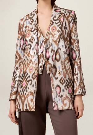 Vrouw draagt een bruin, roze en wit geruit jasje met pailletten en bijpassend vest, gecombineerd met donkerbruine broek, tegen een neutrale achtergrond.