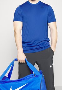 Modré sportovní tričko s krátkými rukávy a kulatým výstřihem, zkombinované s černými kalhotami. Modrá taška přes rameno s bílým logem Nike držena za madlo.
