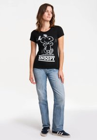 Camiseta de algodón negra con un gráfico blanco de Snoopy, escote redondo y mangas cortas, combinada con unos vaqueros azul claros y zapatillas oscuras.