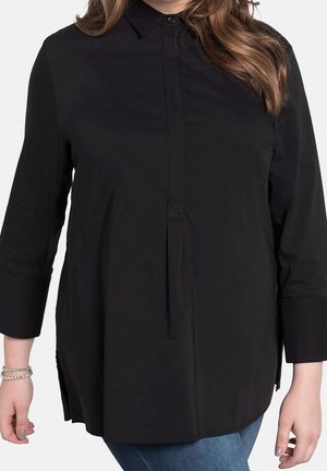 Frau trägt eine lockere schwarze Bluse mit Knöpfen und Dreiviertelärmeln sowie blaue Jeans, sichtbar von den Schultern bis zu den Hüften.