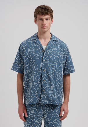 Jonge man die een blauw overhemd met korte mouwen en knopen draagt en een bijpassende broek met witte abstracte lijnpatronen, staand tegen een effen achtergrond.