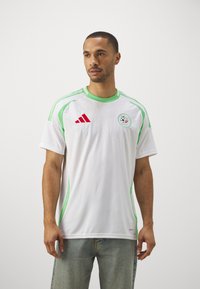Maglietta atletica bianca con accenti verdi, caratterizzata da un collo rotondo, maniche corte, texture intricata e un logo con un motivo calcistico sul petto.