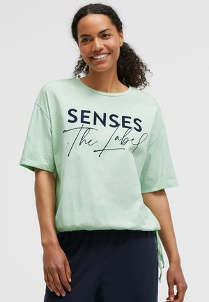 Lächelnde Frau mit lockigem Haar trägt ein hellgrünes T-Shirt mit der Aufschrift "SENSES The Label" und dunkle Hosen vor einfarbigem Hintergrund.