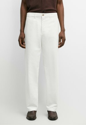 Homme portant un pantalon droit blanc avec une fermeture par bouton et zip, associé à des chaussures marron foncé et un haut marron.
