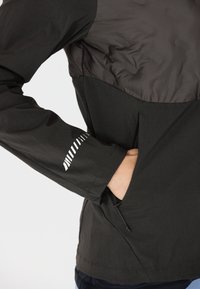 Schwarze Jacke mit einem strukturierten oberen Bereich, einem glatten unteren Bereich und einer Reißverschlusstasche. Reflexive Designdetails am Ärmel.