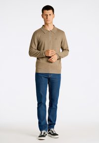 Beige stickad långärmad polo med dragkedja i kragen, bärs med blå jeans och svarta sneakers. Har ribbade muddar och nederkant.