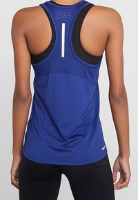 Person med en blå racerback-träningslinne över en svart sport-BH, kombinerat med svarta träningsleggings, vänd bort.