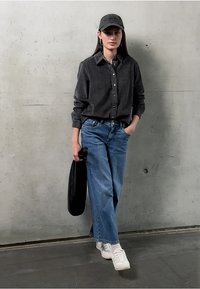 Zwart corduroy button-up shirt, lichtblauwe wijde jeans, witte sneakers en een zwarte pet. Model poseert tegen een betonnen muur.