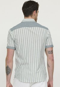 Chemise à manches courtes avec des rayures verticales blanches et bleu pâle, avec un empiècement en chambray et des poignets roulés. Tissu léger, design décontracté.