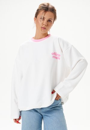 Junge Frau mit hellbraunen Haaren trägt weißen Sweatshirt mit pinkem „offline mode“-Schriftzug und hellblaue Jeans, steht vor weißem Hintergrund.