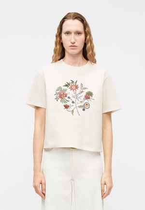 Femme aux longs cheveux ondulés portant une chemise crème à manches courtes avec un motif brodé floral coloré, associée à un pantalon blanc.