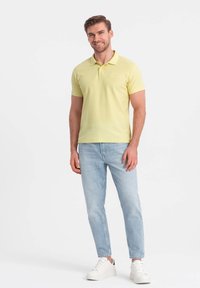 Lys gul polo skjorte med korte ærmer, flad krave og broderet logo; båret med lyseblå jeans og hvide sneakers.
