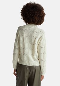 Gruba dzianinowa sweter w kolorze kremowym z wysokim kołnierzem, zdobiony teksturowanym wzorem liści oraz ściągaczami u rękawów, w połączeniu z oliwkowymi spodniami.