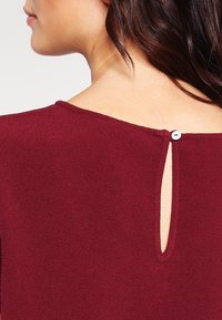 Vêtement bordeaux avec une ouverture en forme de trou de serrure à l'arrière, fini par un petit bouton blanc. Texture lisse et encolure arrondie.