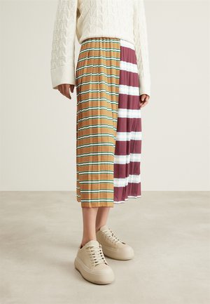 Person iført en midilengde plisseret nederdel med brun-grønne og bordeaux-hvide vandrette striber, cremefarvet sweater og beige platåsneakers.