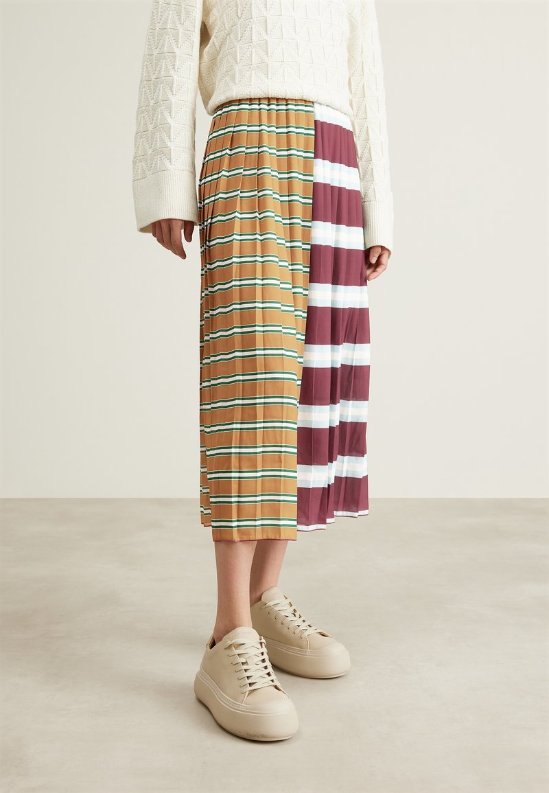 Personne portant une jupe plissée mi-longue avec des rayures horizontales marron-vert et bordeaux-blanc, un pull crème et des baskets beiges à plateforme.