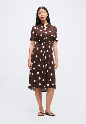 Femme debout dans une robe marron longueur genoux à pois blancs, manches courtes, boutonnée devant, ceinture à la taille et sandales noires.