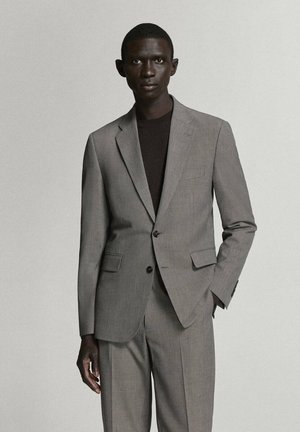 Hombre con un traje gris claro hecho a medida y una camisa negra, de pie con una mano en el bolsillo frente a un fondo liso y claro.