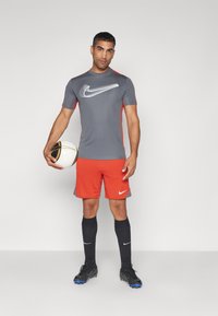 Camiseta gris de Nike con acentos rojos, pantalones cortos naranjas, calcetas negras y botines de fútbol negros. Sostiene un balón de fútbol blanco y dorado.