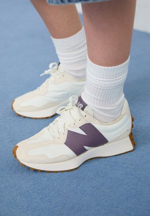 Białe i beżowe trampki z fioletowym logo "N" noszone z białymi prążkowanymi skarpetami za kostkę na niebieskim dywanie.