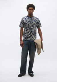 Μπλε σκούρο πουκάμισο με κοντά μανίκια και floral σχέδιο, συνδυασμένο με ανοιχτόχρωμα τζιν και κρατώντας ένα μπεζ πουλόβερ. Μαύρα αθλητικά συμπληρώνουν το σύνολο.