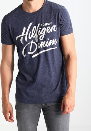 Homme portant un t-shirt bleu marine Tommy Hilfiger Denim et un jean gris, debout devant un fond blanc uni.