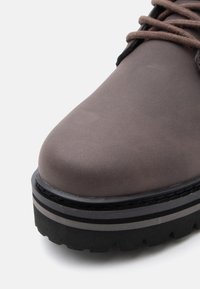 Stivale in pelle marrone con suola nera spessa, caratterizzato da due strisce grigie e lacci marroni su sfondo chiaro.