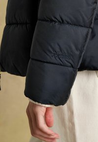 Ruka oblečená v čiernej prešívanej bunde s logom "Joules" na rukáve, odpočíva vedľa svetlých nohavíc.