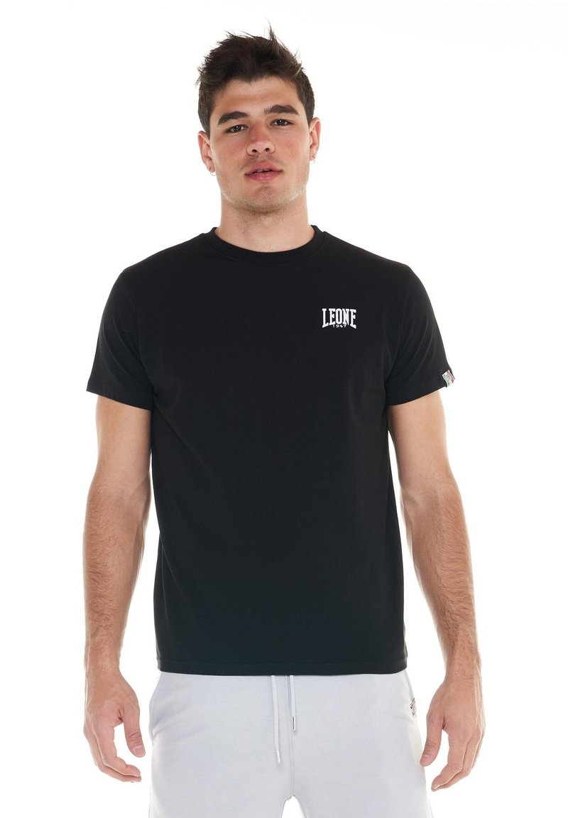 Un homme porte un T-shirt Leone noir avec un petit logo blanc sur la poitrine et un pantalon blanc, sur un fond blanc uni.