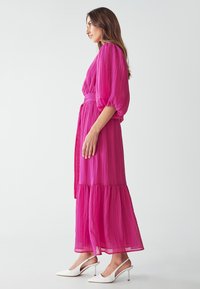 Rosa, gestuftes Maxikleid mit Puffärmeln und gebundenem Taillengurt. Der strukturierte Stoff fällt weich. Kombiniert mit weißen Slingback-Pumps.