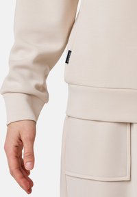 Felpa color crema con vestibilità rilassata, polsini a costine, tasca laterale e piccolo etichetta con logo vicino all'orlo. Morbida al tatto, design minimale.