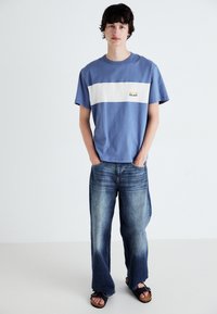 Camiseta de algodón azul con una franja blanca horizontal y un gráfico de sol, combinada con pantalones vaqueros anchos de un azul desvanecido y sandalias negras.