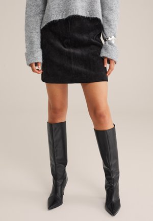 Mini-jupe en velours côtelé noir associée à un pull en laine grise et à des bottes en cuir noir taille genou avec des pointes.