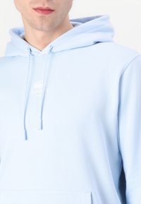 Šviesiai mėlynas džemperis su gobtuvu, pagamintas iš minkštos medžiagos, turi kengūros kišenę, raištelių gobtuvą ir siuvinėtą Lacoste logotipą krūtinėje.