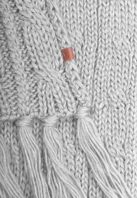 Tissu tricoté gris clair avec de grosses torsades et des extrémités effilochées, avec une petite étiquette marron intégrée au design.