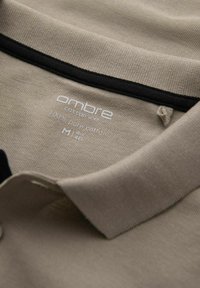 Krémové polo tričko vyrobené ze 100% bavlny. Má černý výstřih a štítek s informacemi o značce a velikosti.