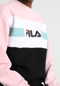 Różowa, biała i czarna bluza z poziomymi paskami; zawiera jasnoniebieski pas oraz logo FILA w czerni. Materiał bawełniany, okrągły dekolt.