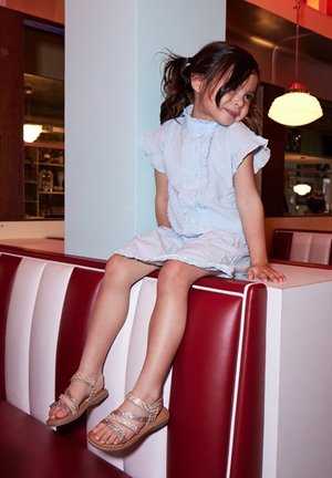 Himmelblaues Kleid mit Rüschenaermeln, sitzt an einem rot-weißen Tisch. Trägt beige Sandalen mit dekorativen Riemen und einer strukturierten Sohle.