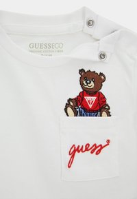 Vit ekologisk bomull T-shirt med en broderad björn som bär en röd skjorta och blå jeans, med en framsidig ficka och röd "guess" broderi.