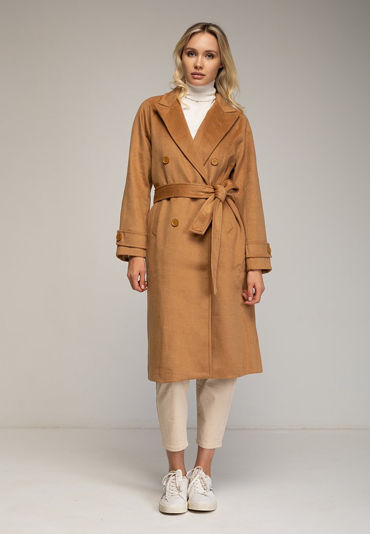 Cappotto in misto cammello con design doppiopetto abbottonato, vita con cintura, ampi rever e maniche lunghe. Indossato sopra pantaloni leggeri e sneaker.
