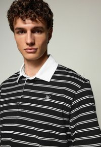 Sort-hvid stribet polo-shirt med hvid krave og et lille broderet logo på brystet. Blødt, struktureret stof.