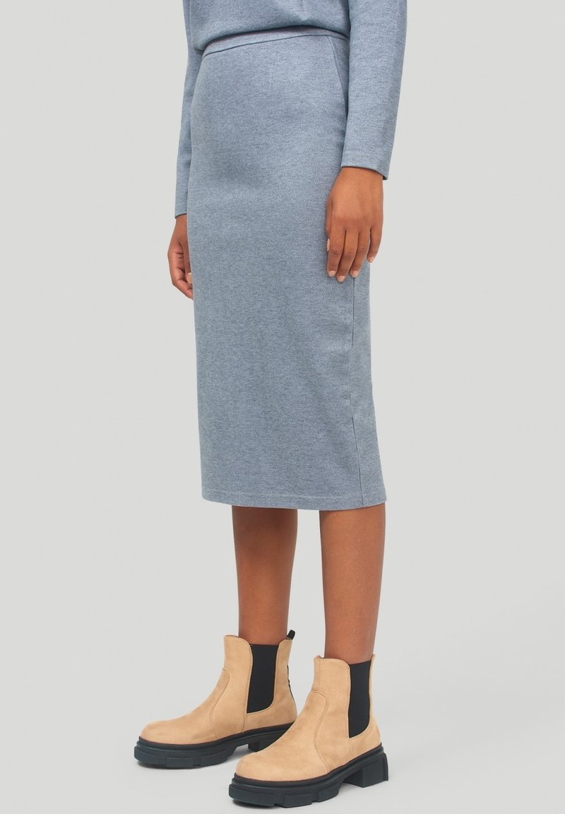 Personne portant une jupe crayon bleue longueur genoux et des bottines beige avec des panneaux latéraux élastiques noirs, debout sur un fond uni.