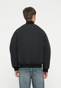 Sort bomberjakke med rund krave, polstret tekstur og elastiske manchetter. Modellen har lys blå jeans på med synlige sømme.