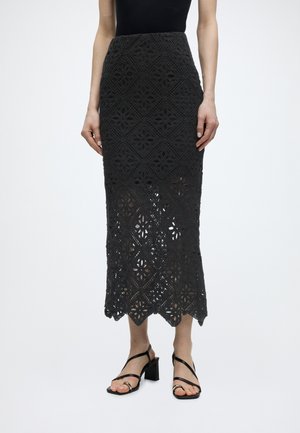 Frau trägt einen schwarzen, hoch taillierten Häkel-Midi-Rock mit floralen Muster und gewelltem Saum, kombiniert mit schwarzen Sandalen mit Riemchen und Blockabsatz.